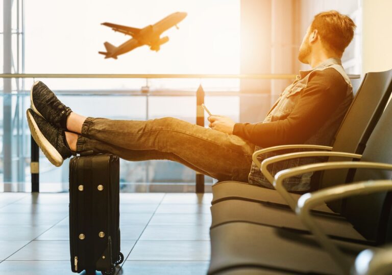 Mężczyzna czeka na lotniksu opierając nogi o walizkę oraz przyglądając się odlatującym samolotom