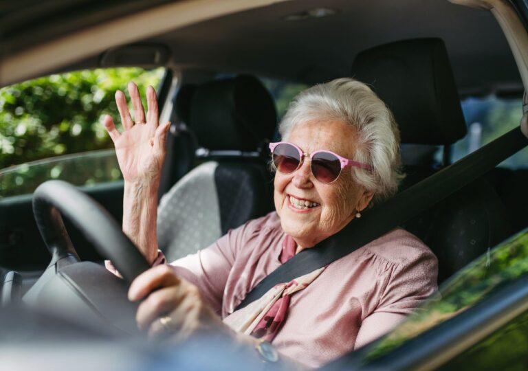 Starsza kobieta w okularach przeciwsłonecznych macha uśmiechnięta siedząc w samochodzie