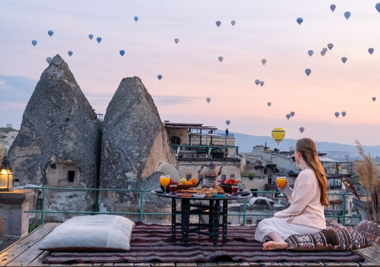 Na tle formacji skalnych Kapadocji kobieta ogląda balony pijąc herbatę
